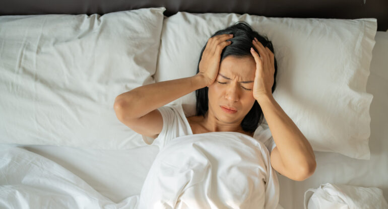 Woman in bed with hands on head experiencing chronic migraine pain