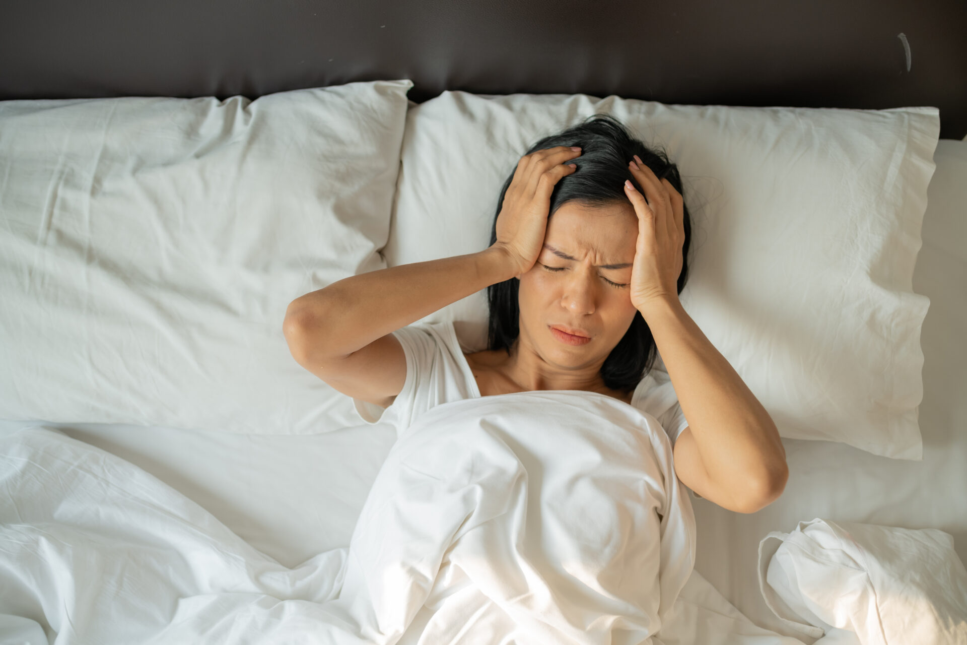 Woman in bed with hands on head experiencing chronic migraine pain