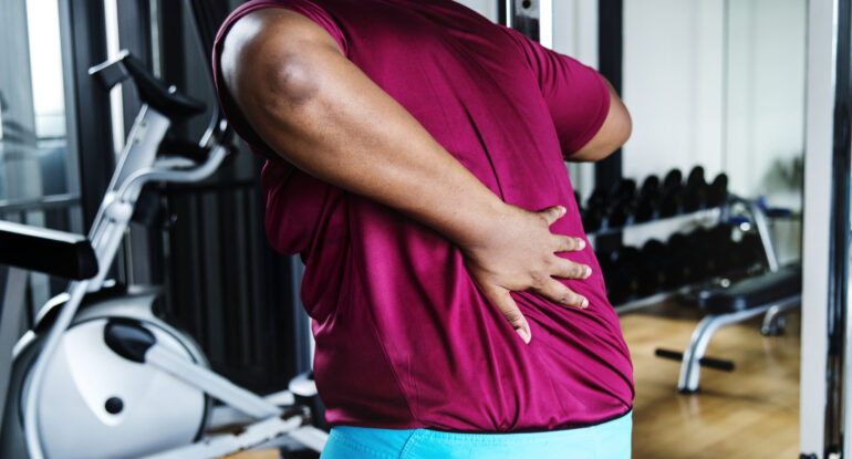Man with lower back pain from a gym injury, holding his back near workout equipment