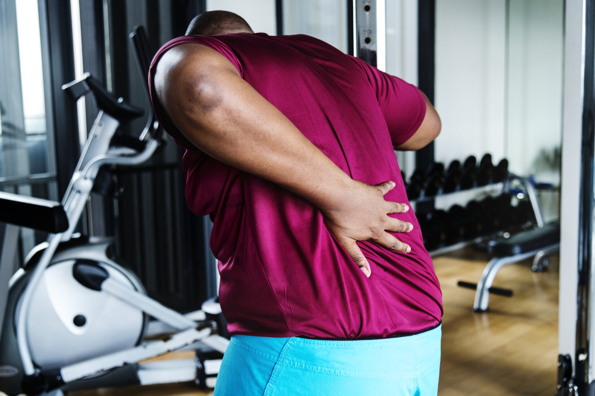 Man with lower back pain from a gym injury, holding his back near workout equipment